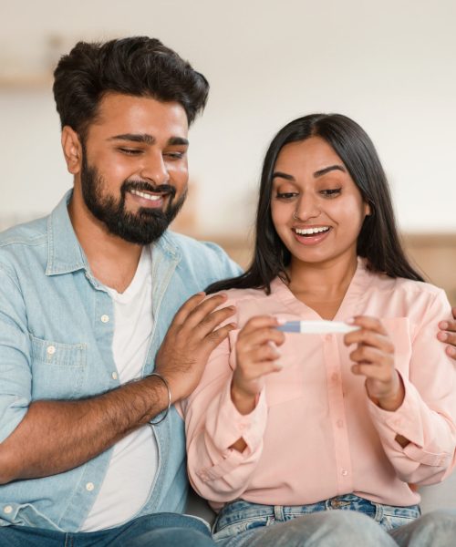 Happy young indian spouses looking at positive pregnancy test, excited of becoming parents, sitting on couch at home Happy young indian spouses looking at positive pregnancy test, excited of becoming parents, sitting together on couch at home. Loving hindu couple awaiting baby