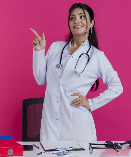 smiling young female doctor wearing medical robe and stethoscope standing behind desk with medical tools keeping hand on waist looking and pointing at side isolated on pink background